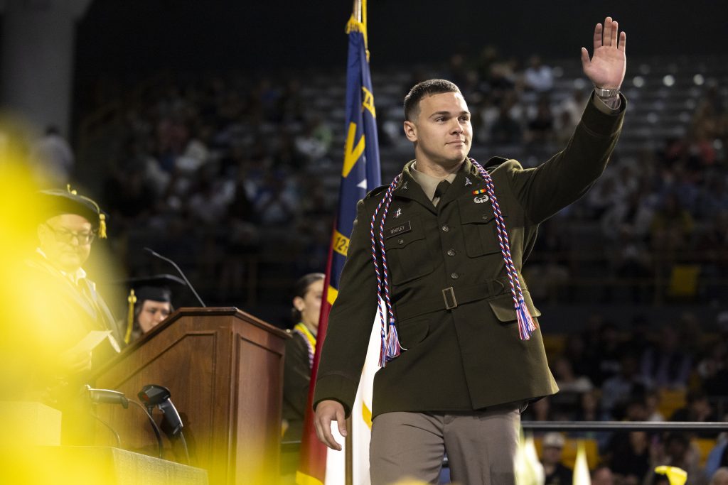 A student walking across a graduation stage at Appalachian State University in a military uniform, waving to the audience with graduation cords around his neck.