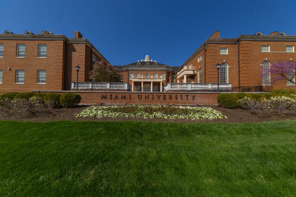A photo of Miami University's campus, low brown buildings behind a sprawling green lawn.