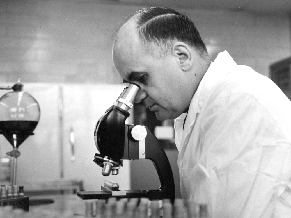 A black and white photo of a man peering into a microscope.
