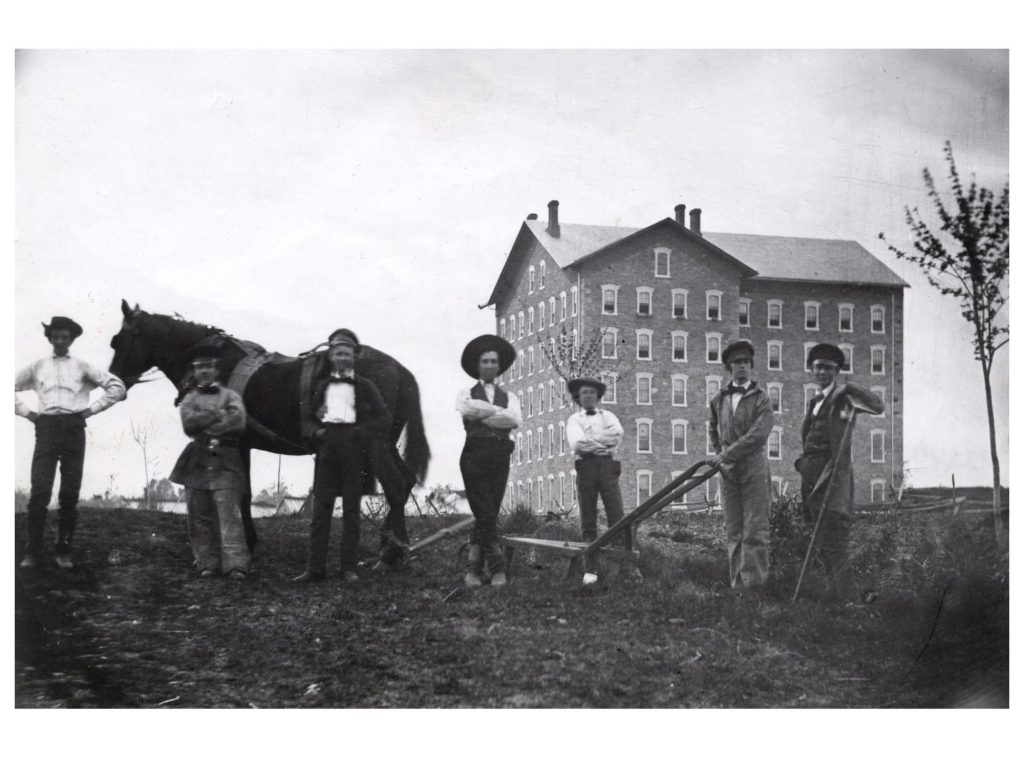A black and white photo of a group of farmers and a horse in front of a building.