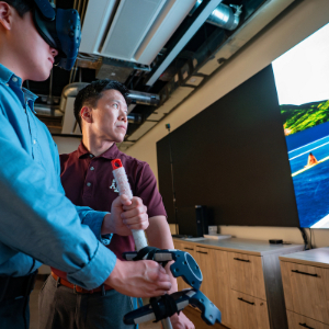 A photo of two people, one in a VR headset, standing in front of a television simulating a road test.