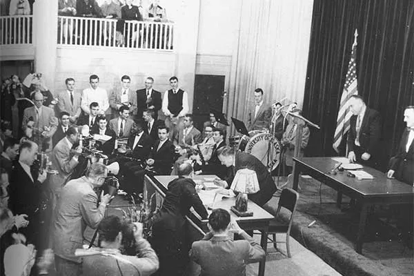 A black and white photo of a constitutional convention in Alaska. The room is full of men in suits and a an is bent over a document, seemingly signing it, near the front of the room.