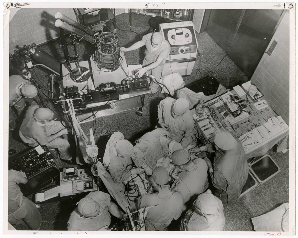 A black and white aerial photo of an operating room full of doctors and nurses in scrubs operating on a patient.