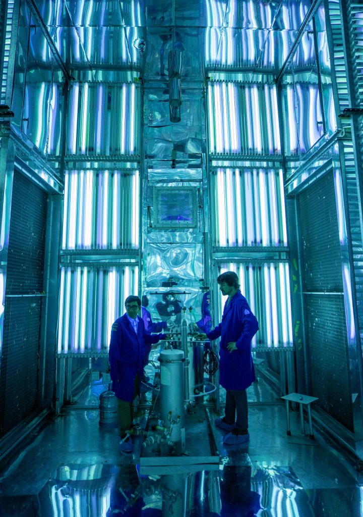 A photo of two scientists in blue jackets in an atmospheric chamber for air pollution research. The chamber is blue and white with lights glowing throughout.