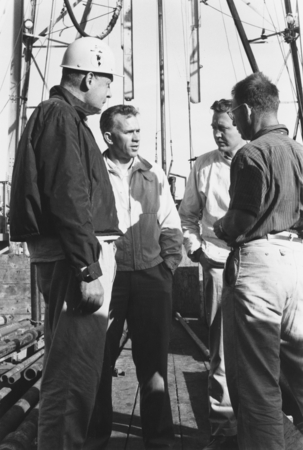 A black and white photo of four men speaking intensely on a boat.